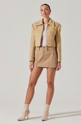 A woman is wearing a cropped, textured, beige jacket with a collar and long sleeves. She has on a white top underneath, a beige mini skirt with front seams and belt loops, and beige high-heeled ankle boots with pointed toes.